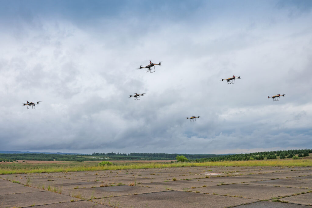 Skupina transportních dronů Malloy letí nad letištní plochou pod zataženou oblohou.