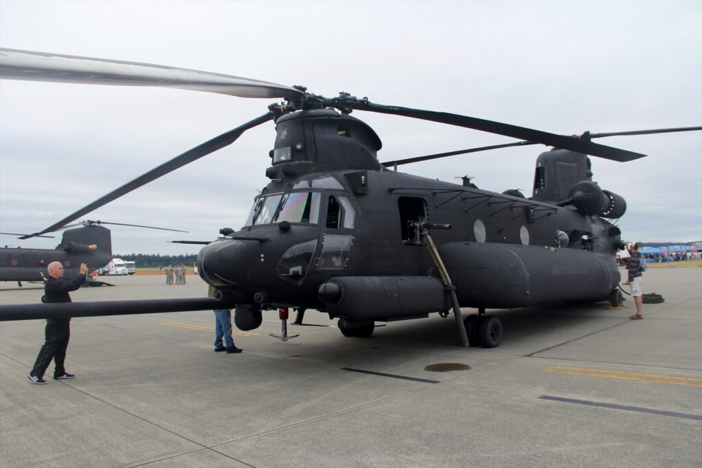 Tmavý vrtulník Chinook se dvěma rotory a dlouhým nástavcem před přídí stojí mezi návštěvníky na letišti.