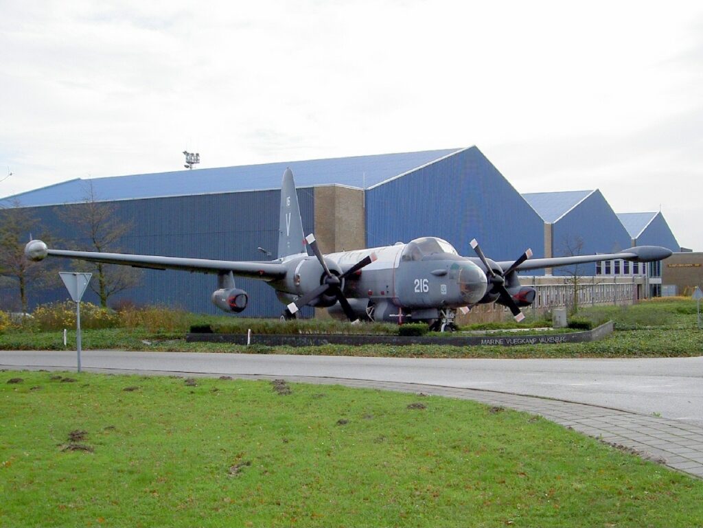 Šedý letoun P2V-7 Neptune s číslem 216 vystavený na travnaté ploše před hangárem.