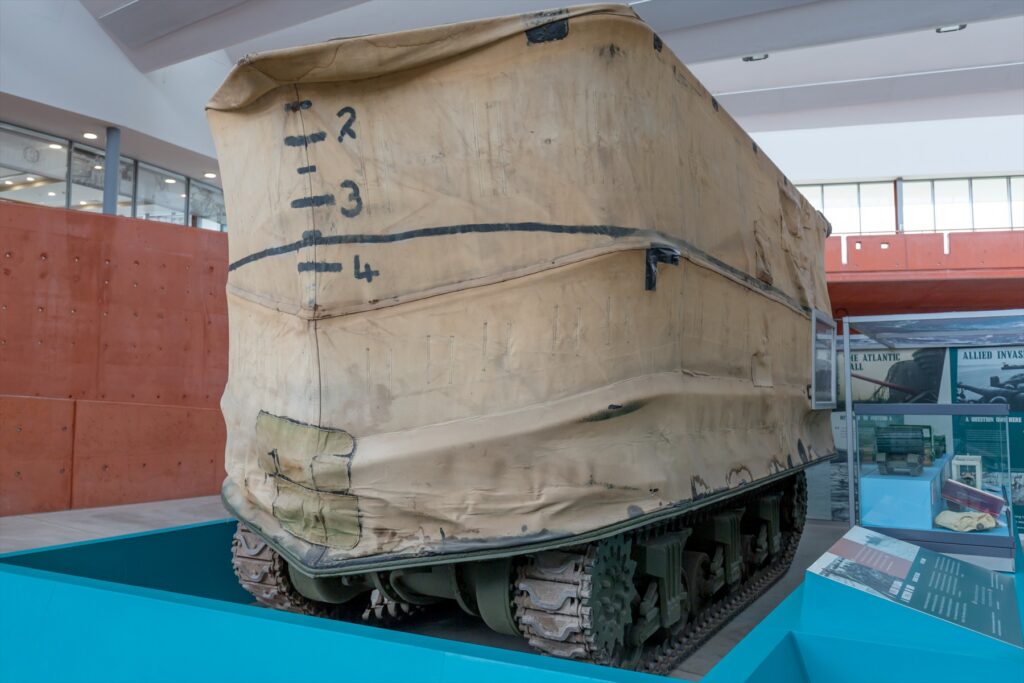 Muzejní tank Sherman DD zakrytý vysokou plátěnou stěnou, pod níž jsou vidět pásy