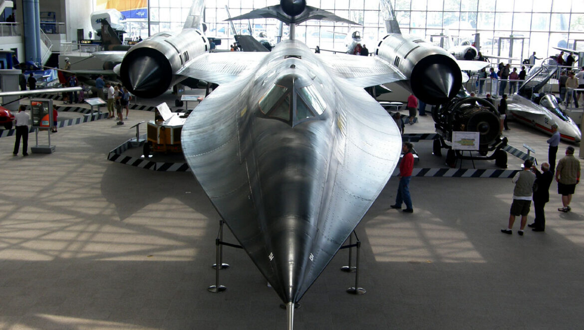 Čelní pohled na letoun Lockheed M-21 s kovovým potahem a menším černým dronem D-21 nad zadní částí trupu v muzejní hale.