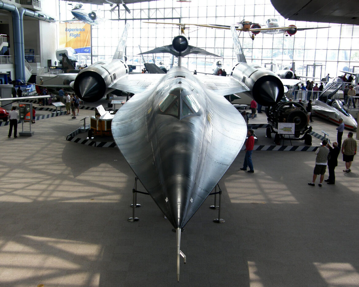 Čelní pohled na letoun Lockheed M-21 s kovovým potahem a menším černým dronem D-21 nad zadní částí trupu v muzejní hale.
