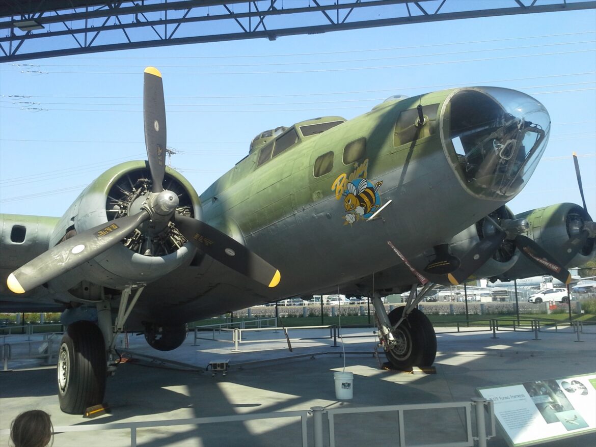 Zelený bombardér Boeing B-17F s prosklenou přídí a vrtulovými motory pod muzejním přístřeškem.