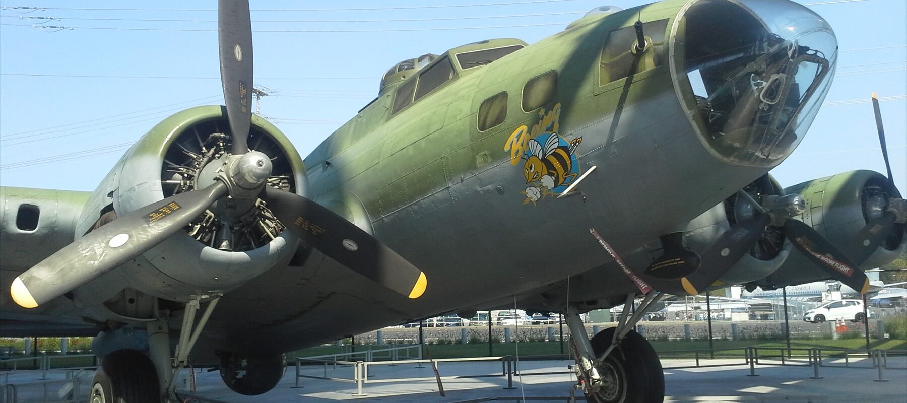 Zelený bombardér Boeing B-17F s prosklenou přídí a vrtulovými motory pod muzejním přístřeškem.
