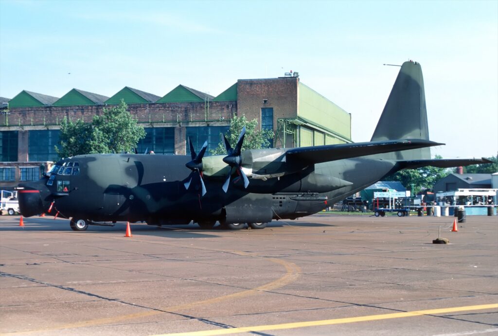 Kamuflovaný čtyřmotorový letoun MC-130E stojící na letištní ploše před hangáry.