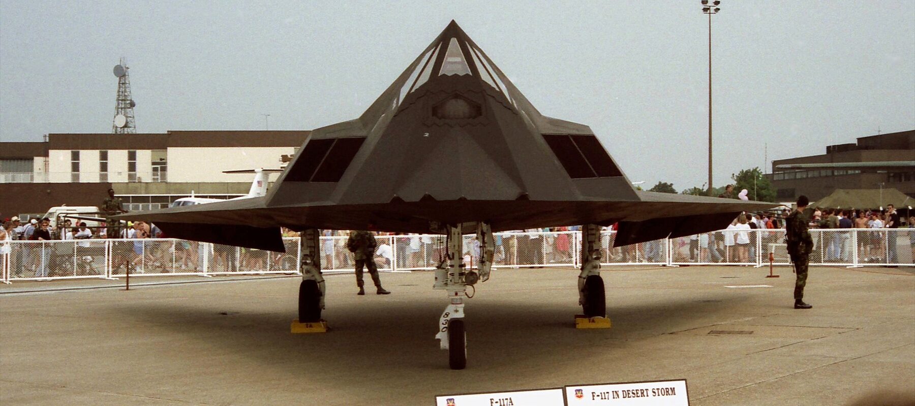 Černý F-117A na podvozku při čelním pohledu, za zábranami stojí návštěvníci.