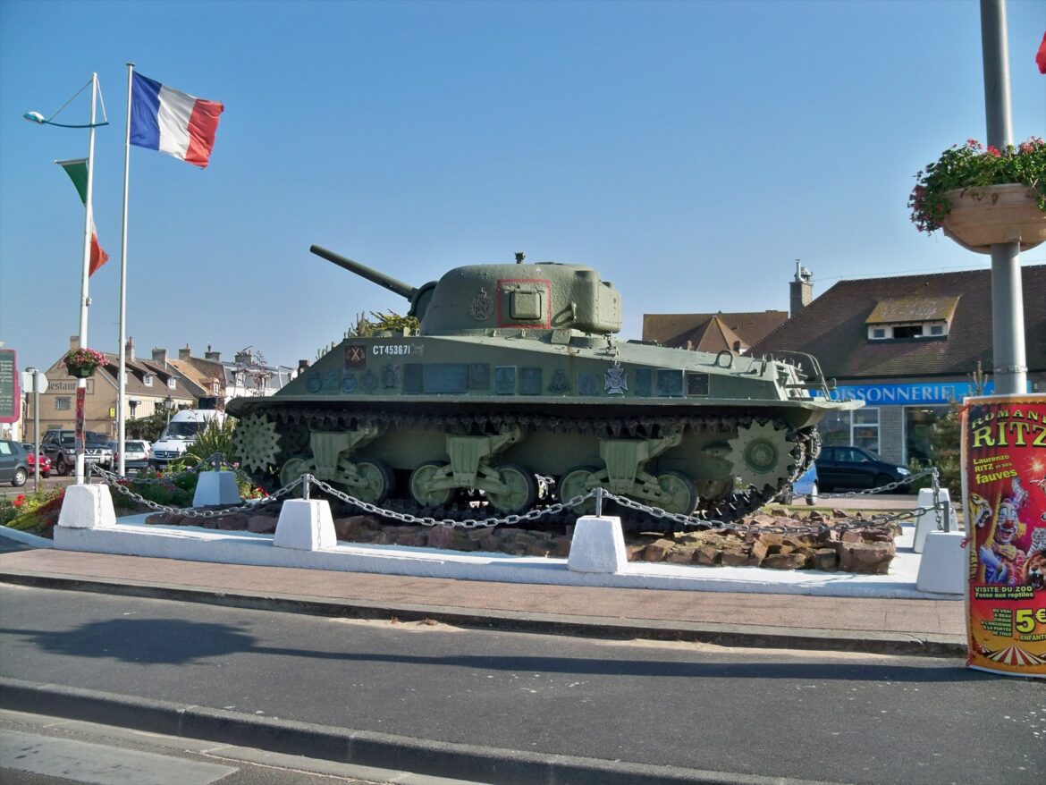 Zelený tank Sherman DD na kamenném podstavci u ulice a vlajkových stožárů