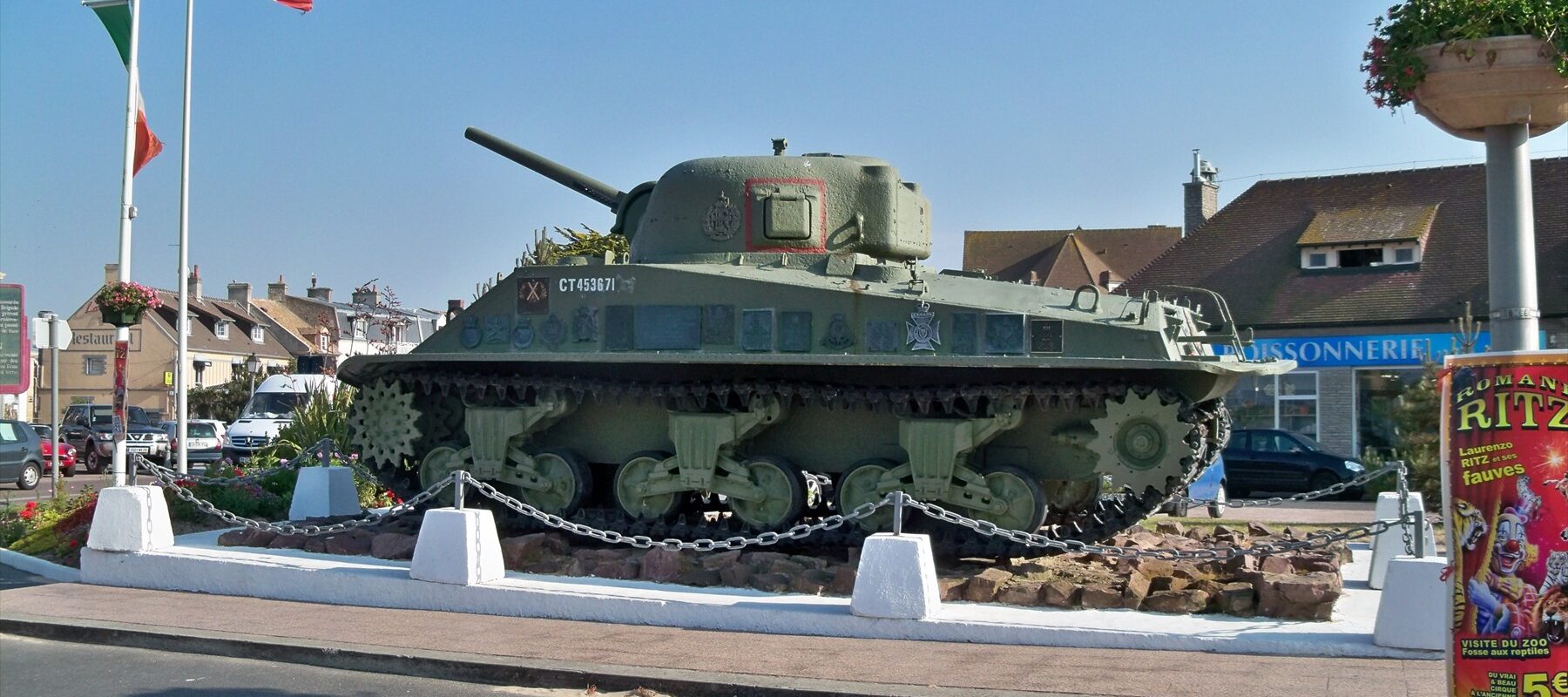 Zelený tank Sherman DD na kamenném podstavci u ulice a vlajkových stožárů
