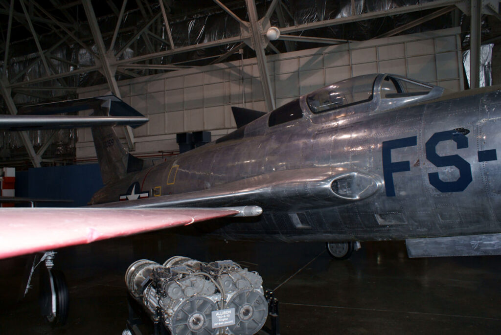 Bok XF-84H s kabinou, trojúhelníkovou plochou za ní a ocasem ve tvaru T; v popředí muzejní motor.