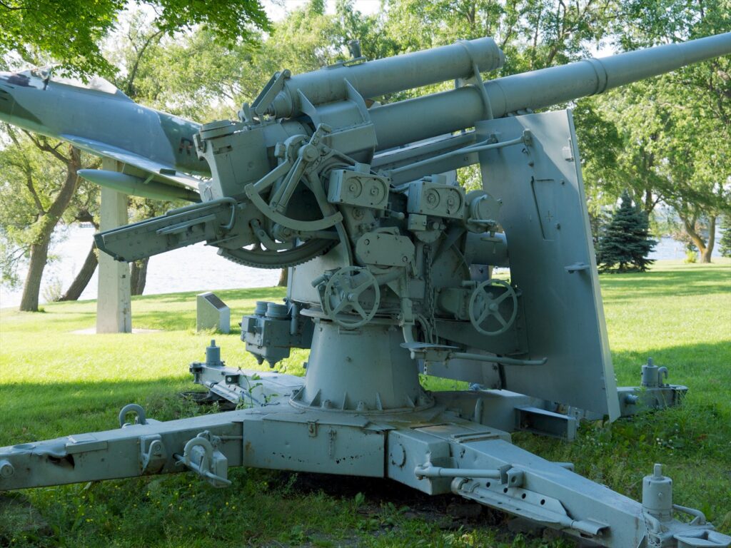 Šedý kanon Flak 37 s dlouhou hlavní a ochranným štítem v parku u vody.