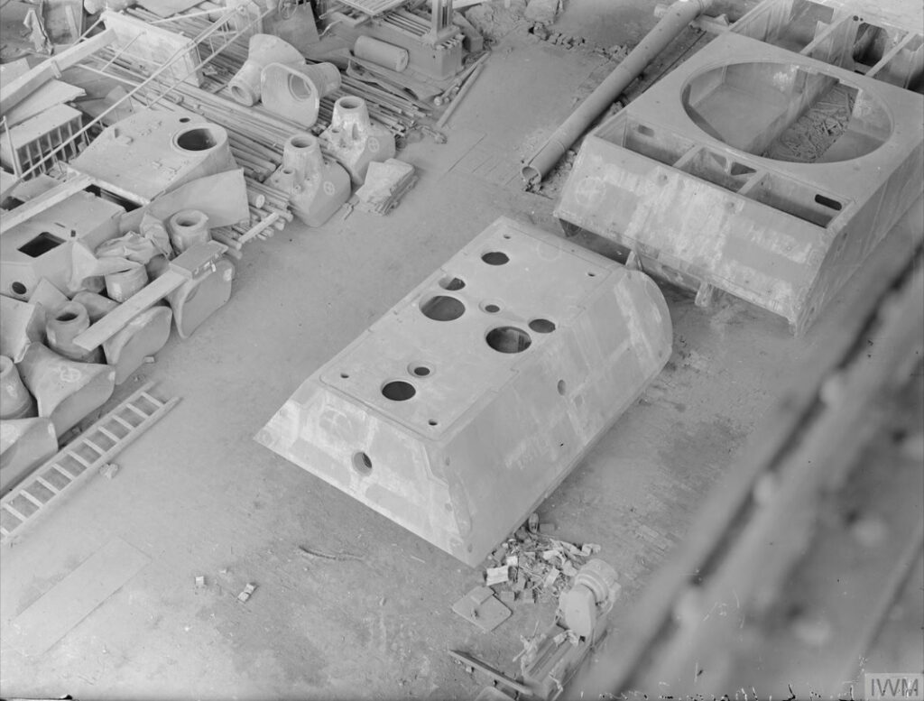Dobový černobílý snímek z roku 1945: nedokončená věž a korba tanku Maus mezi rozpracovanými díly v obsazené německé továrně