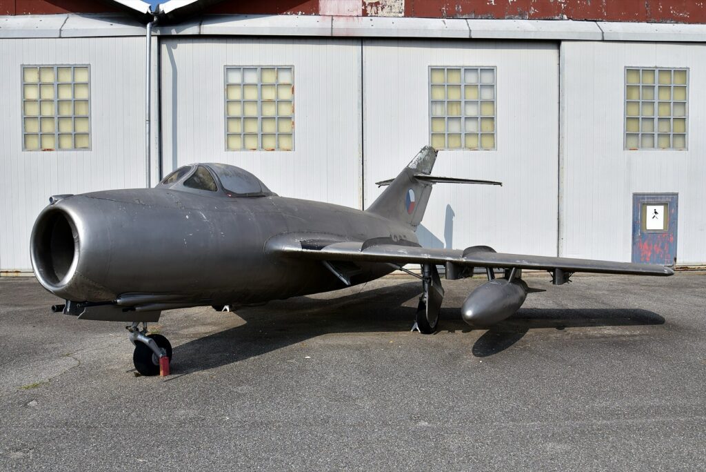 Podzvukový stíhací letoun MiG-15 v československých barvách vystavený u hangáru, boční pohled