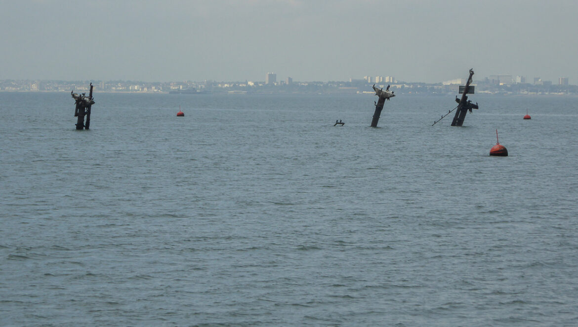 Tři nakloněné stožáry vraku SS Richard Montgomery trčící z hladiny ústí Temže, v pozadí obrys pobřeží