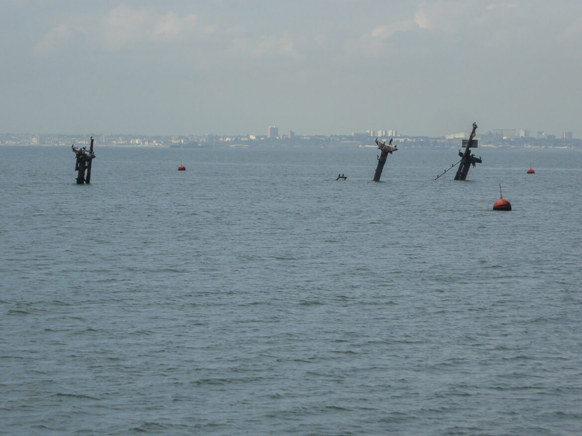 Tři nakloněné stožáry vraku SS Richard Montgomery trčící z hladiny ústí Temže, v pozadí obrys pobřeží