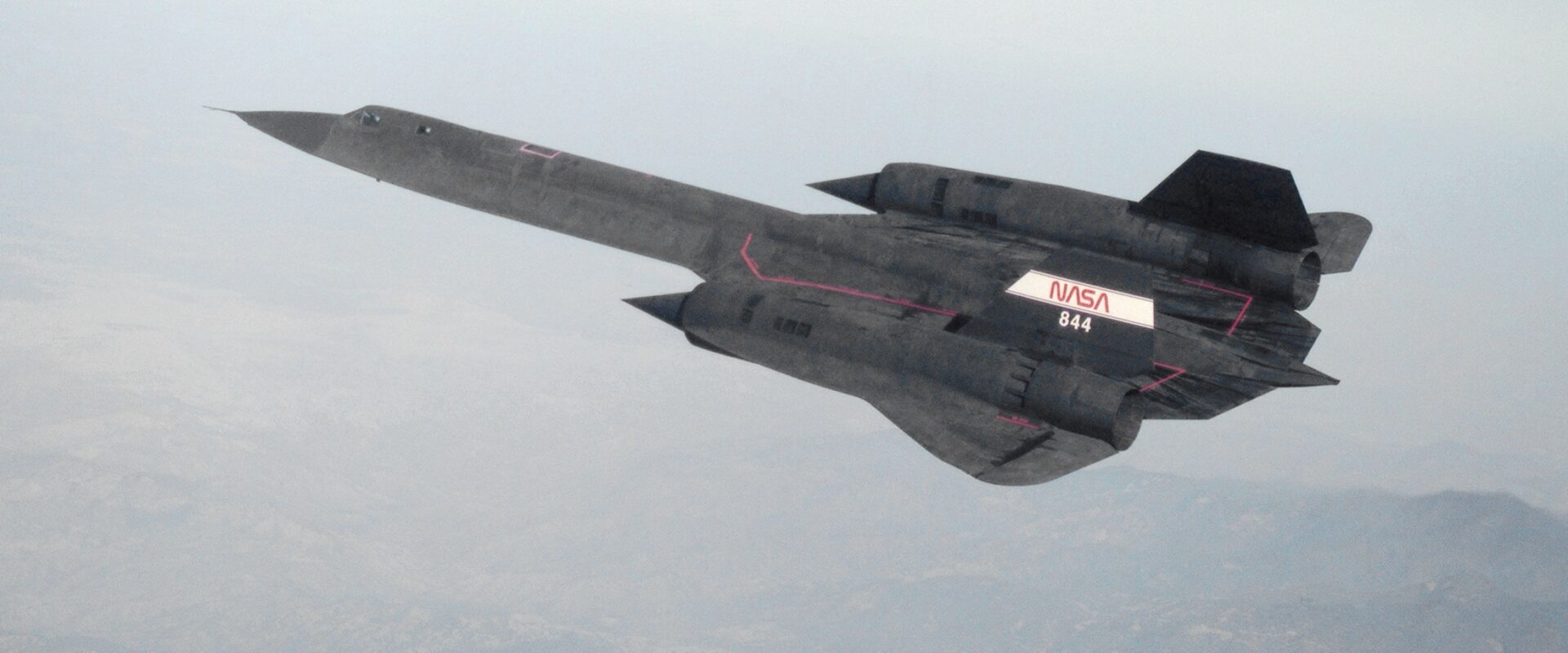 Průzkumný letoun Lockheed SR-71 Blackbird v letu nad horami, pohled shora s dlouhými trupovými přechody a delta křídlem