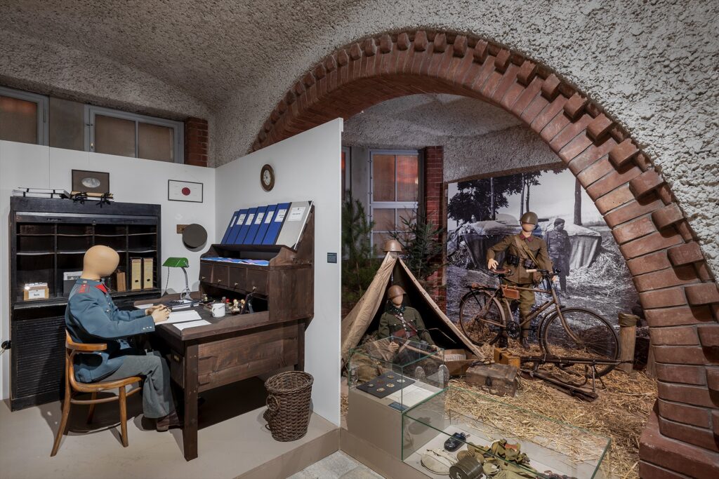 Muzejní diorama polního ležení s vojenským stanem, jízdním kolem a přilbou vzor 32