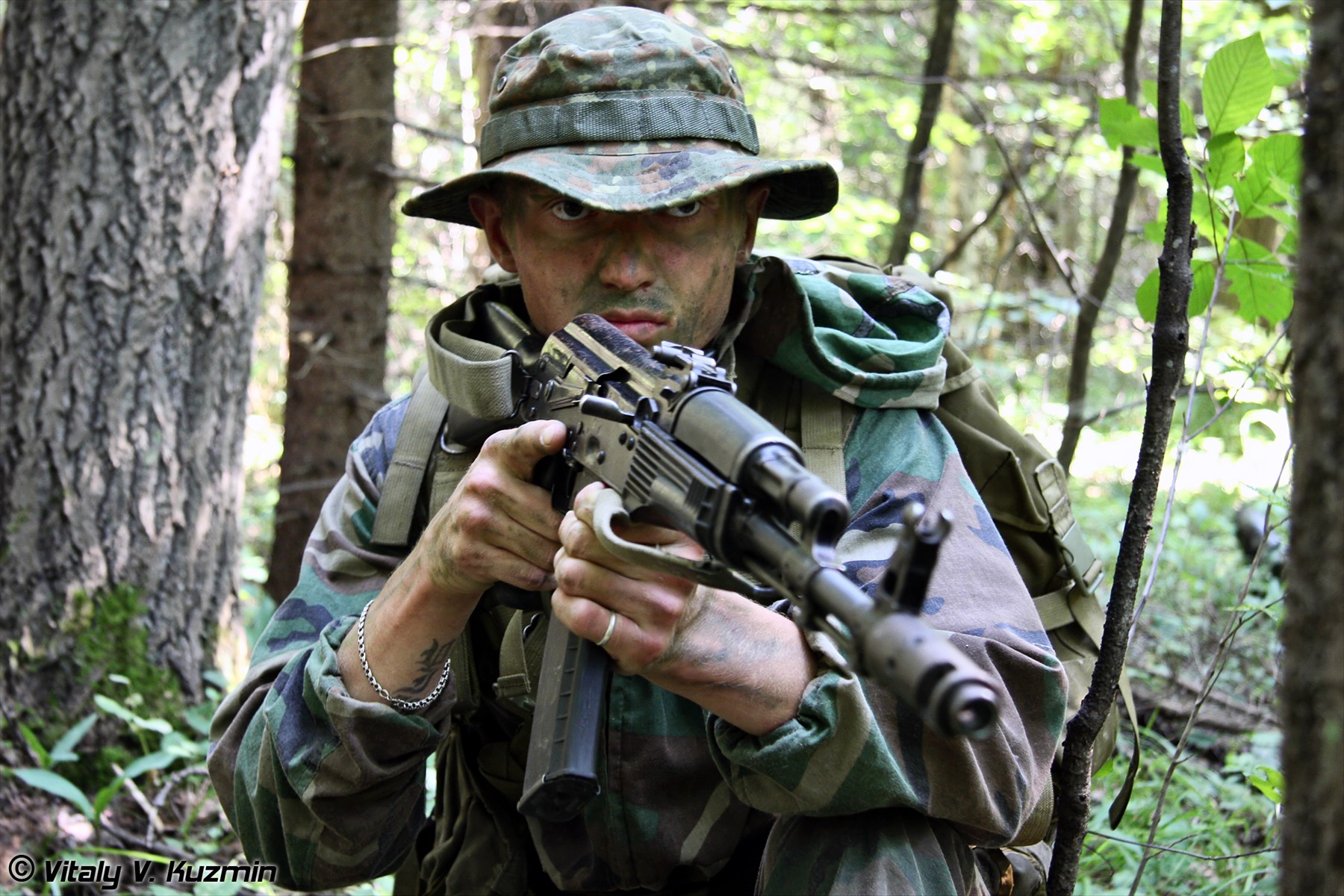 Střelec v maskovací uniformě mířící z útočné pušky AK-74 s oranžovým plastovým zásobníkem a úsťovou brzdou.