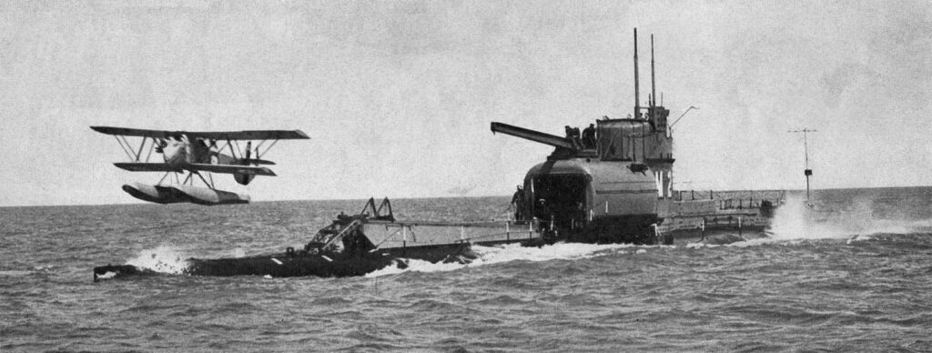 Hydroplán Parnall Peto ve vzduchu před přídí ponorky HMS M2 s otevřeným hangárem.
