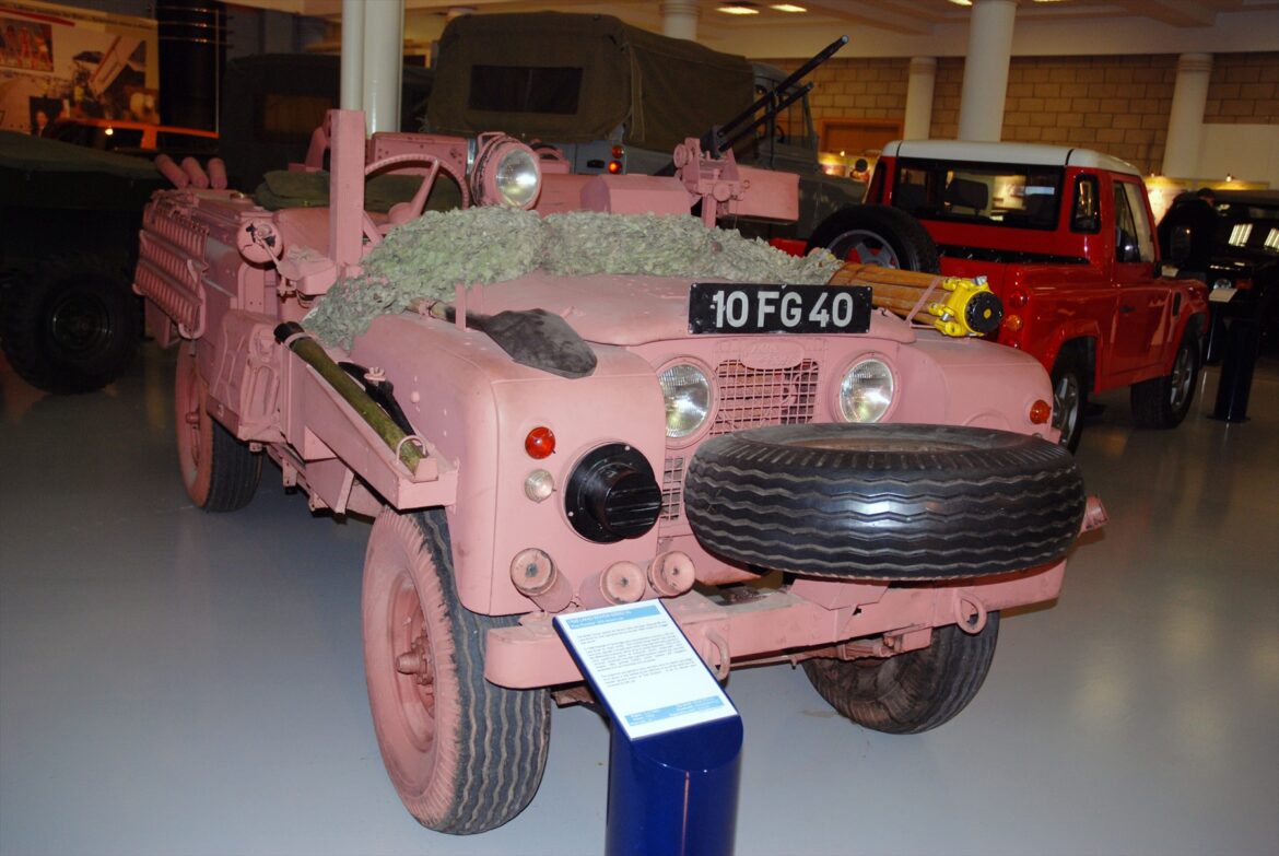 Růžový otevřený Land Rover s označením 10 FG 40 a rezervním kolem vpředu v muzeu.