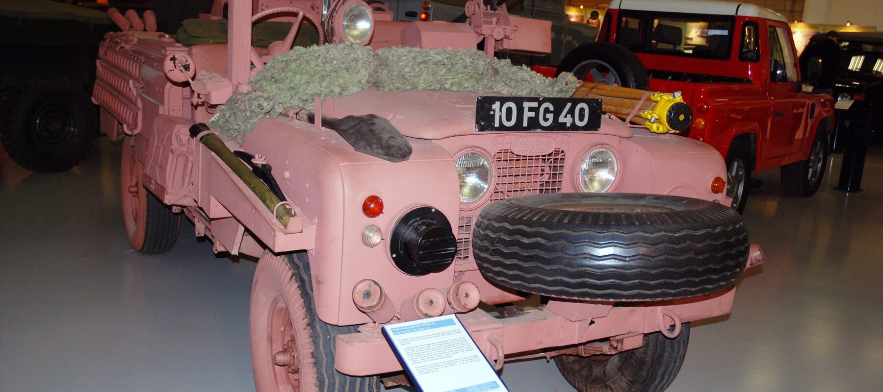Růžový otevřený Land Rover s označením 10 FG 40 a rezervním kolem vpředu v muzeu.