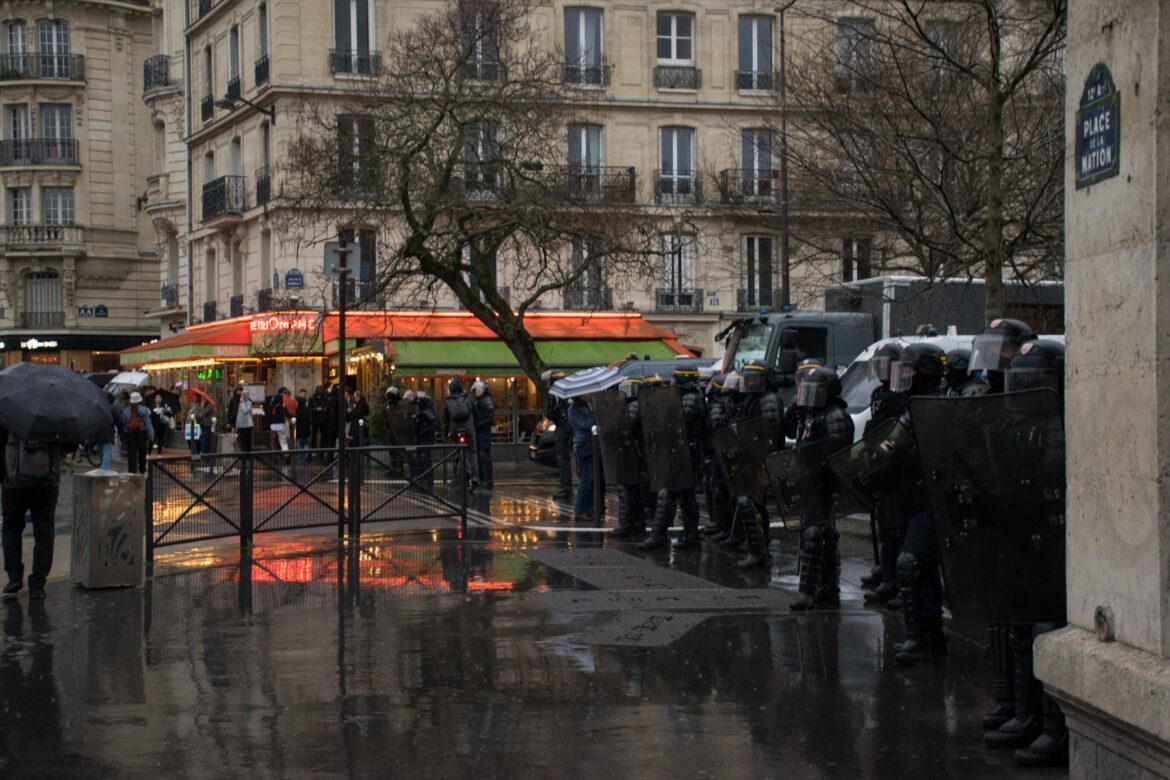 Řada policistů v přilbách s hledím a se štíty na deštivém náměstí Place de la Nation.
