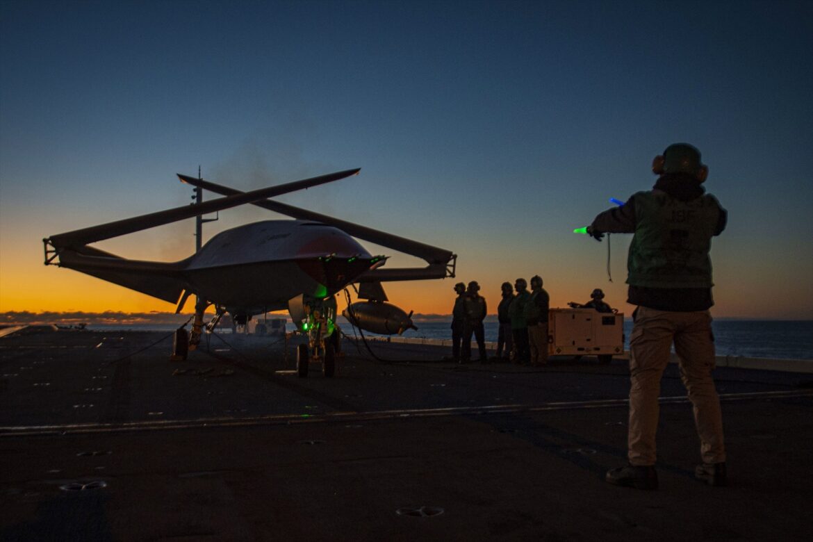 Bezpilotní MQ-25 se složenými křídly a palubní personál na letadlové lodi za soumraku.