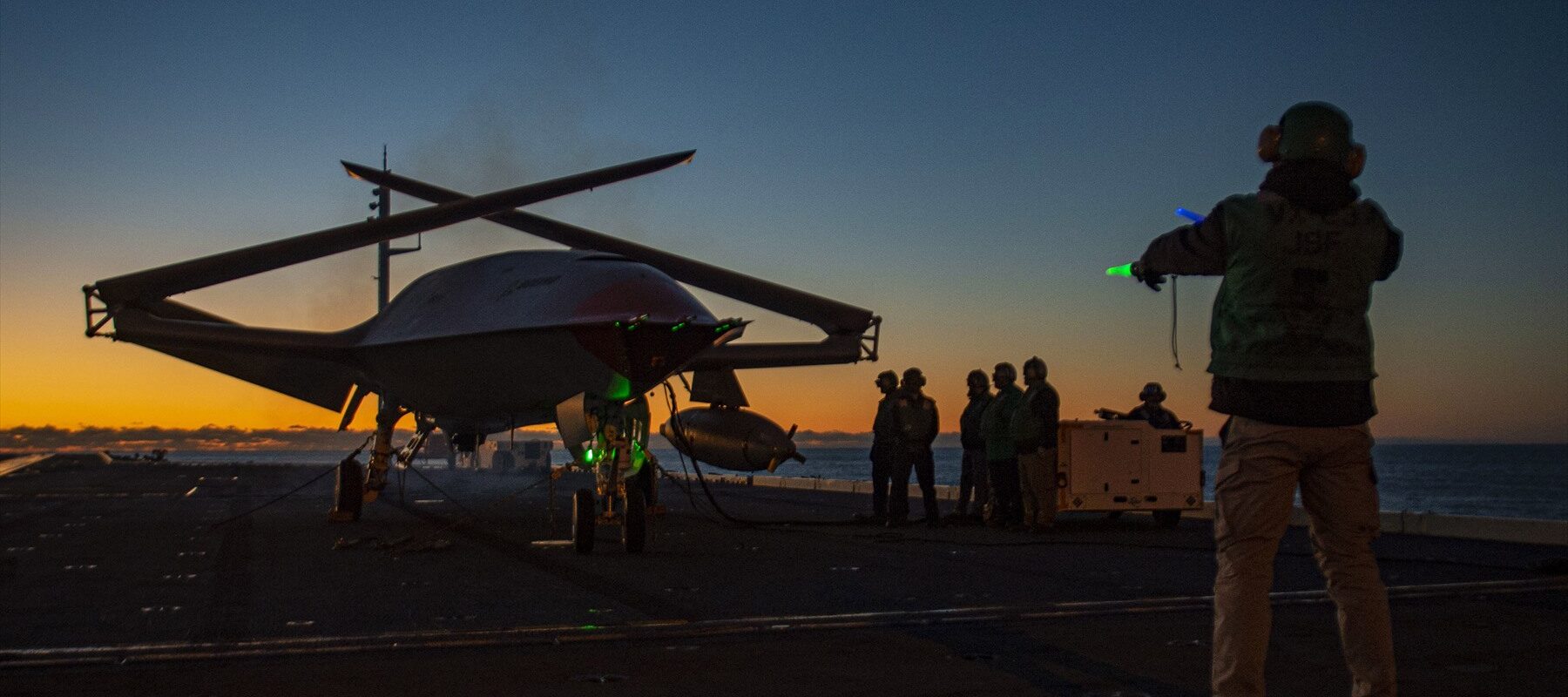 Bezpilotní MQ-25 se složenými křídly a palubní personál na letadlové lodi za soumraku.