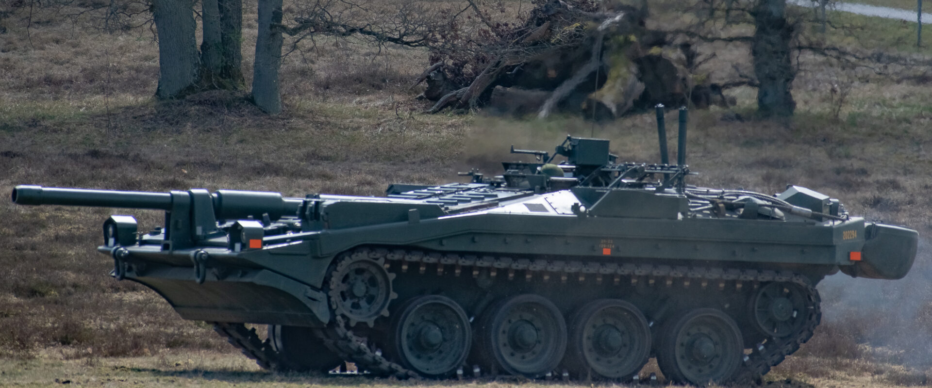 Pásový tank Strv 103 bez otočné věže projíždějící travnatým vojenským prostorem.